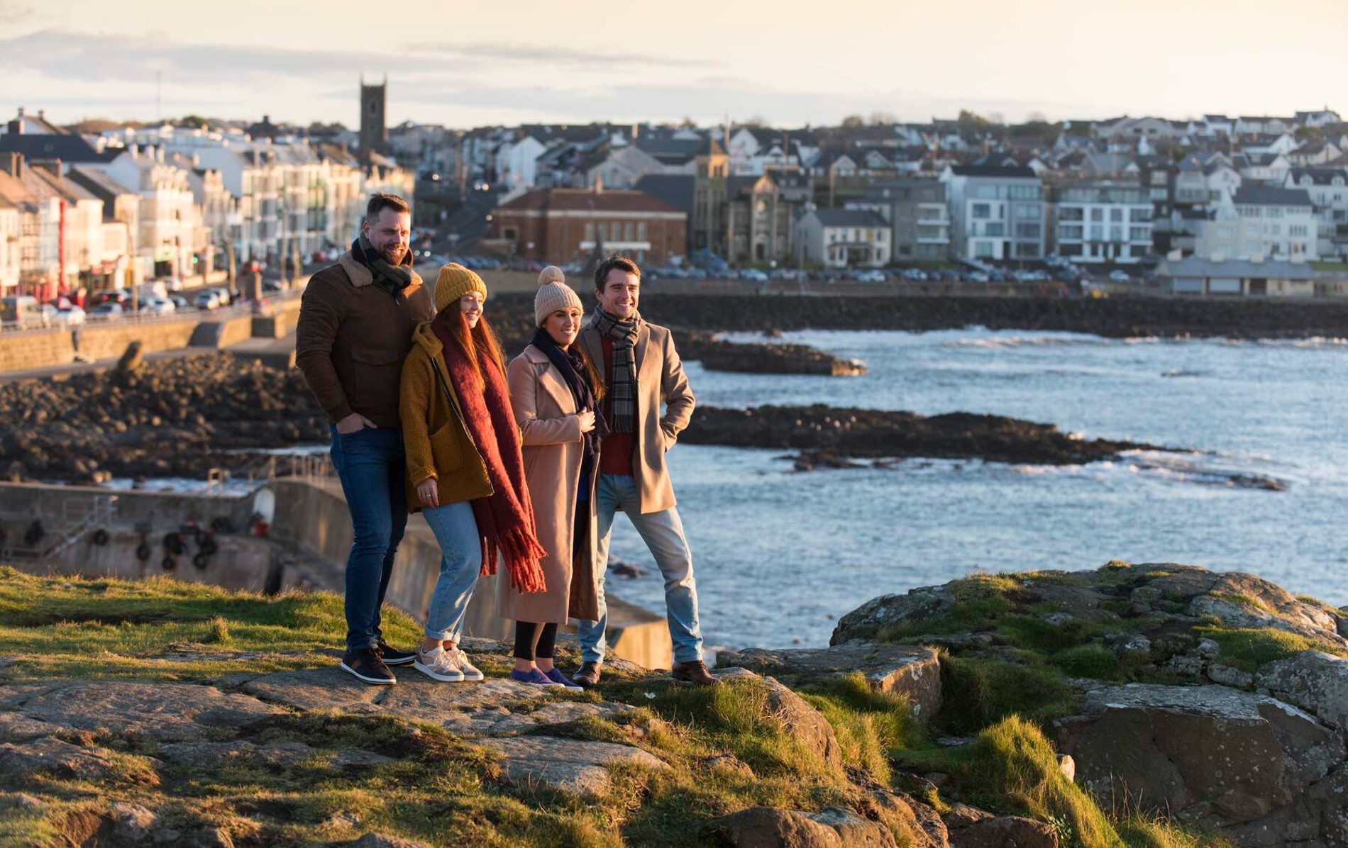 Four friends in coats and hats stand on cliffs, overlooking the sea and town houses of Portstewart.