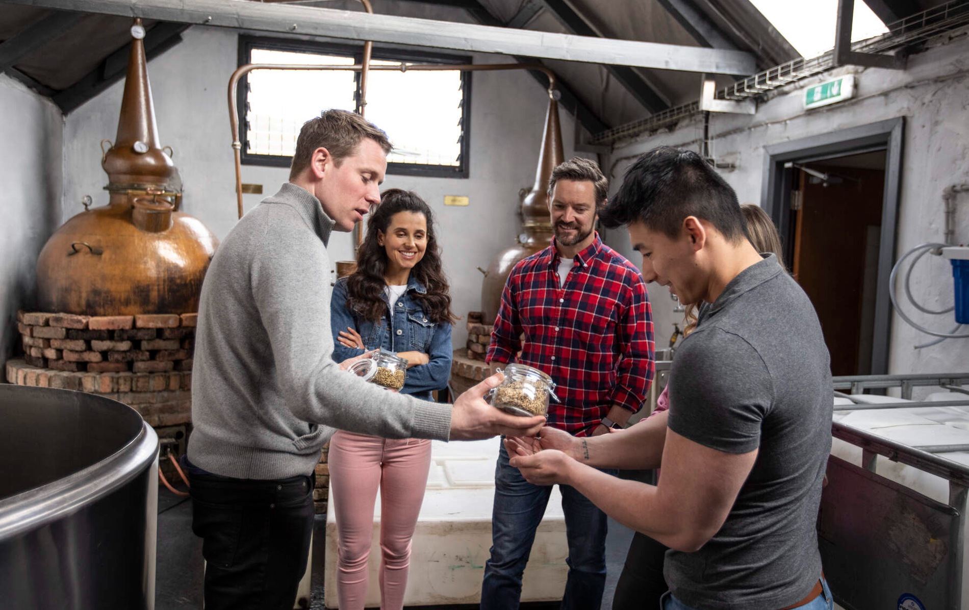 Visitors at Killowen Distillery in County Down handle jars of grains beside copper stills during a guided tour.