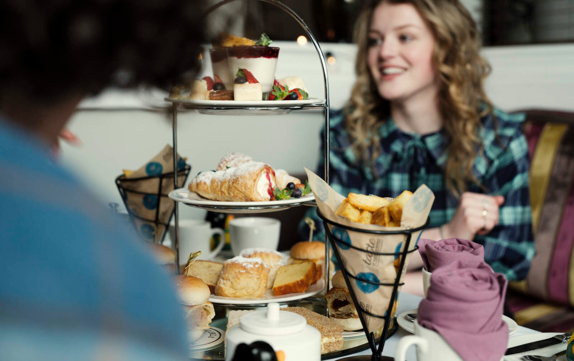 Tiered stand of scones, cakes, chips, and pastries as friends chat happily during themed afternoon tea.