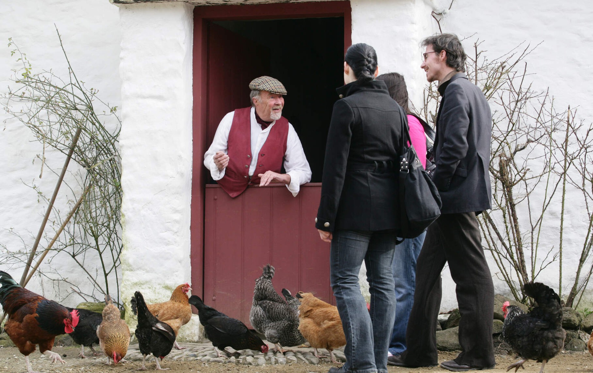 Guide in waistcoat and cap greets visitors at a farmhouse door as hens scatter at their feet.
