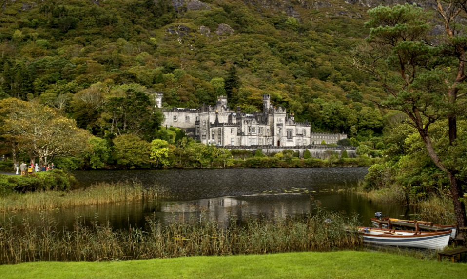 Abbaye de Kylemore se reflétant dans un lac immobile au pied d’une colline boisée, avec une barque en bois au bord de l’eau.