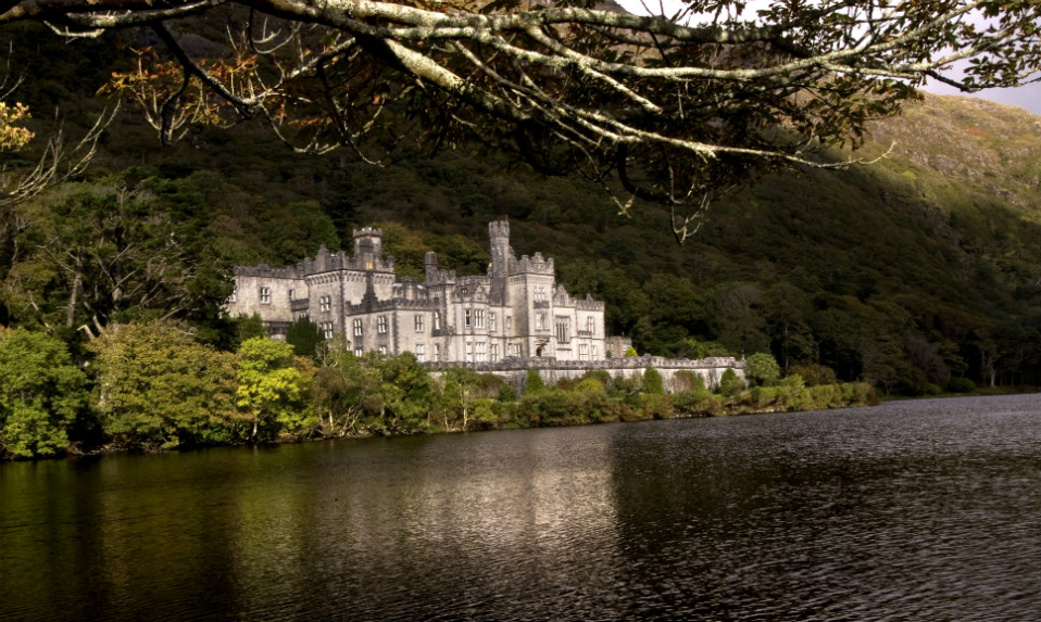 Neo-Gothic Kylemore Abbey reflected in a tranquil lake, surrounded by trees beneath the slopes of Connemara hills.