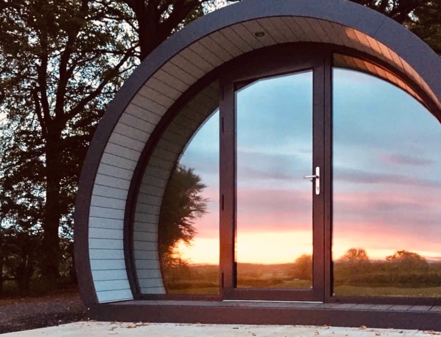 Un glamping pod curvo alla Thornfield Farm, nella contea di Antrim, riflette l'alba rosa e il paesaggio rurale.