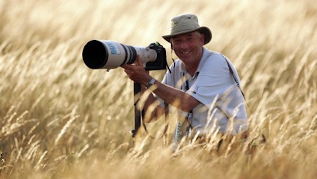 Der Golf-Fotograf David Cannon kniet im hohen Gras mit einer Kamera mit langem Objektiv und lächelt, während er sein Ziel ins Visier nimmt.