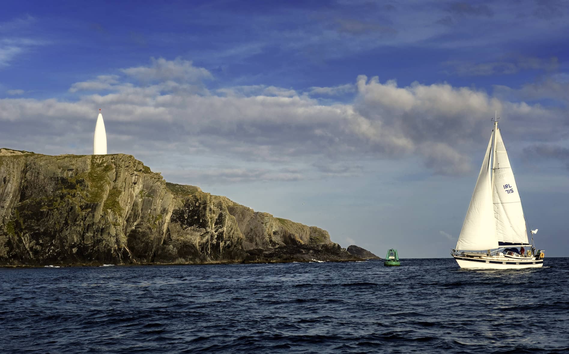 Una barca a vela scivola lungo le ripide scogliere con un faro bianco lungo la costa della contea di Cork.