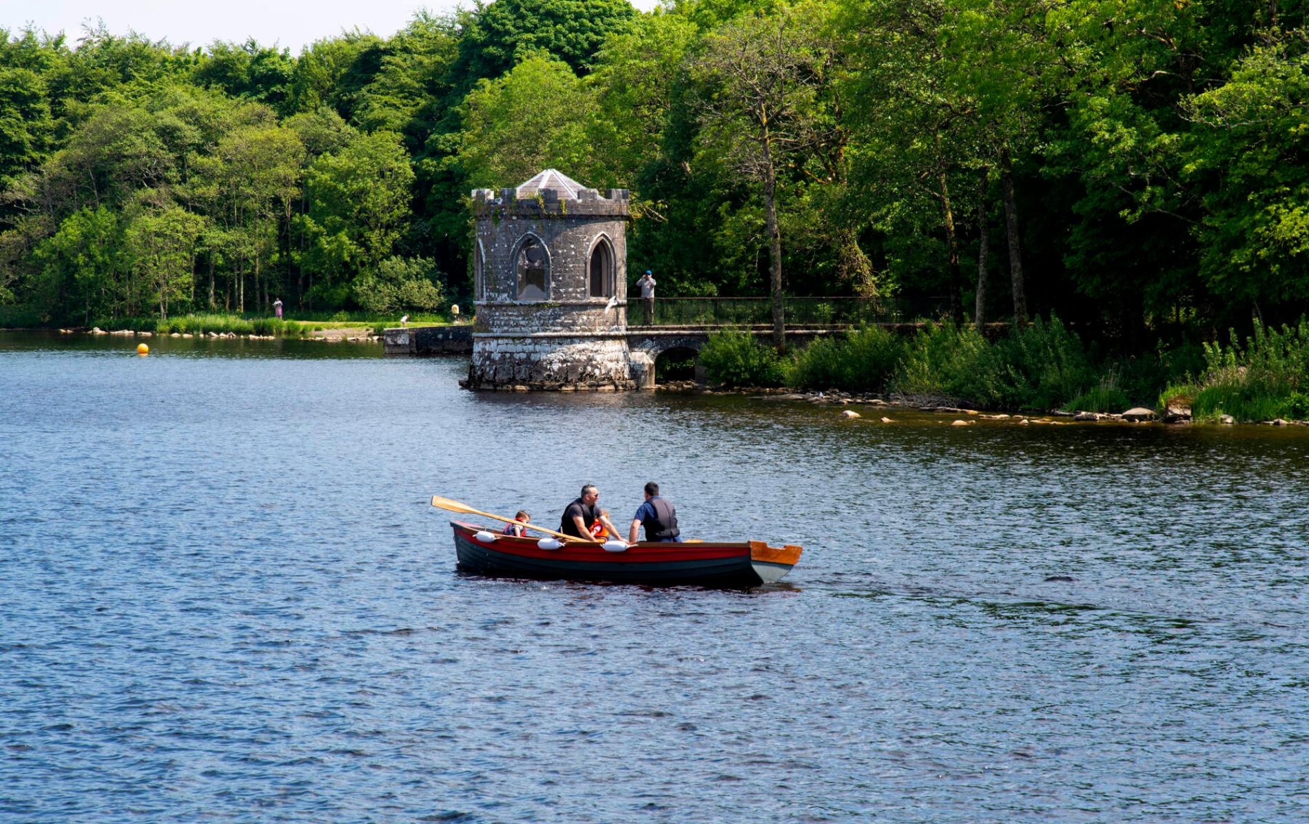 Een roeiboot met drie personen op Lough Key, county Roscommon, in de buurt van een klein stenen botenhuisje, omgeven door bomen.