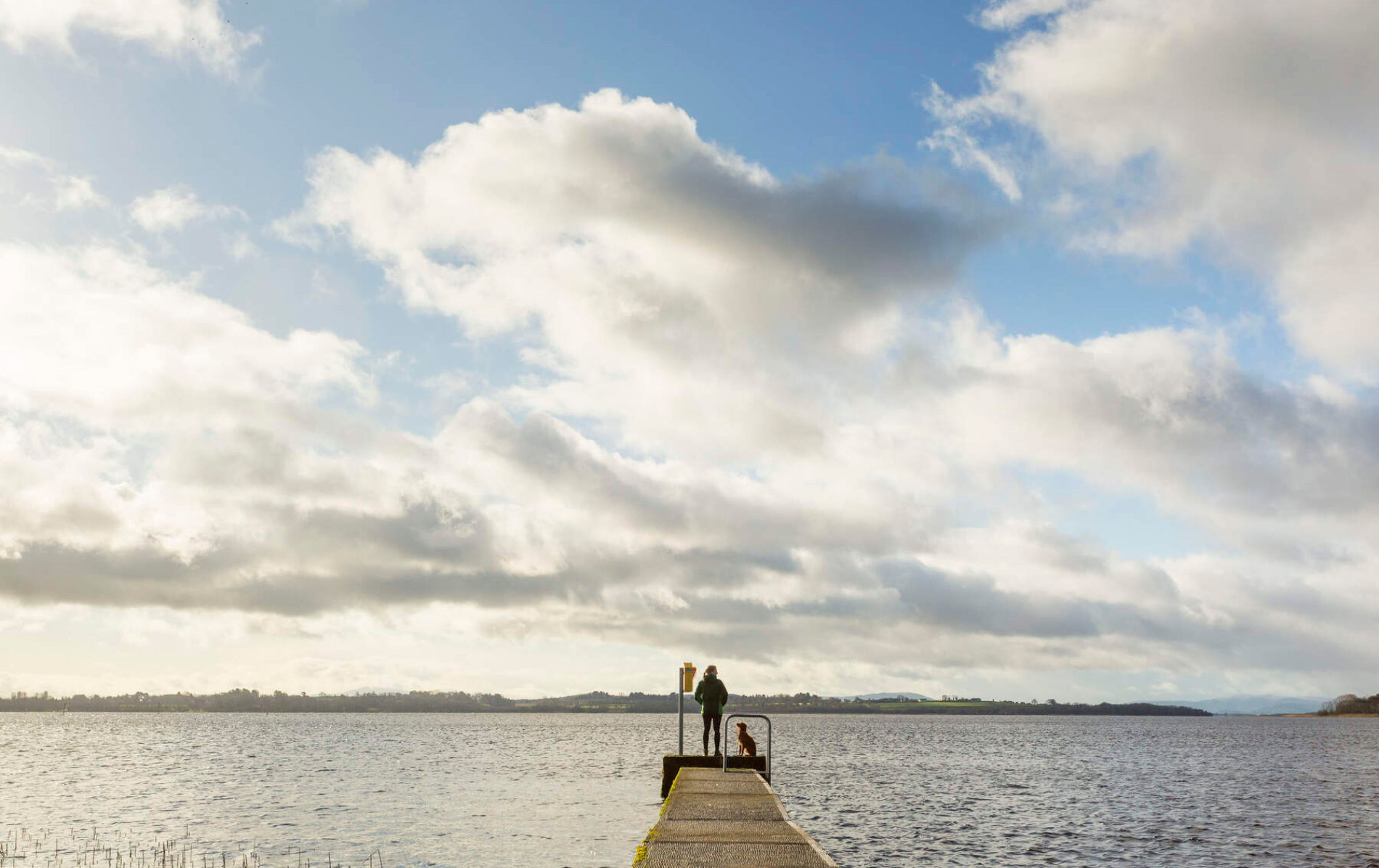 Man en hond staan aan het einde van een steiger met uitzicht op Lough Derg in county Galway.