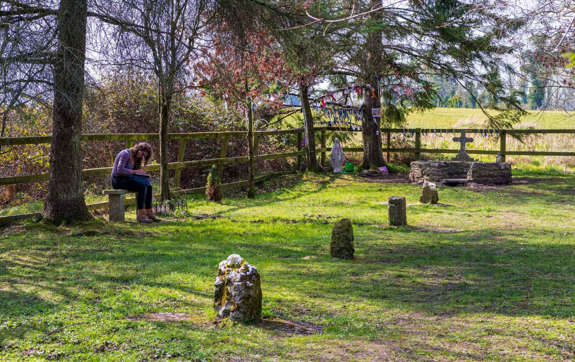Sacred well beneath tall trees with statues and ribbons as a woman reflects on a stone bench.