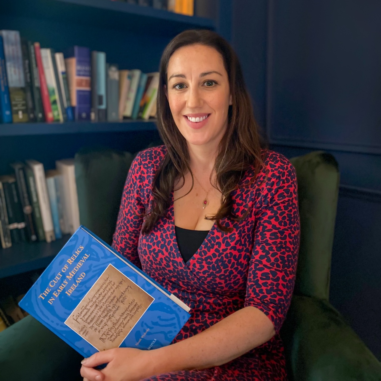 Historian Dr Niamh Wycherley smiles holding her book, seated in a study of Irish saint relics.