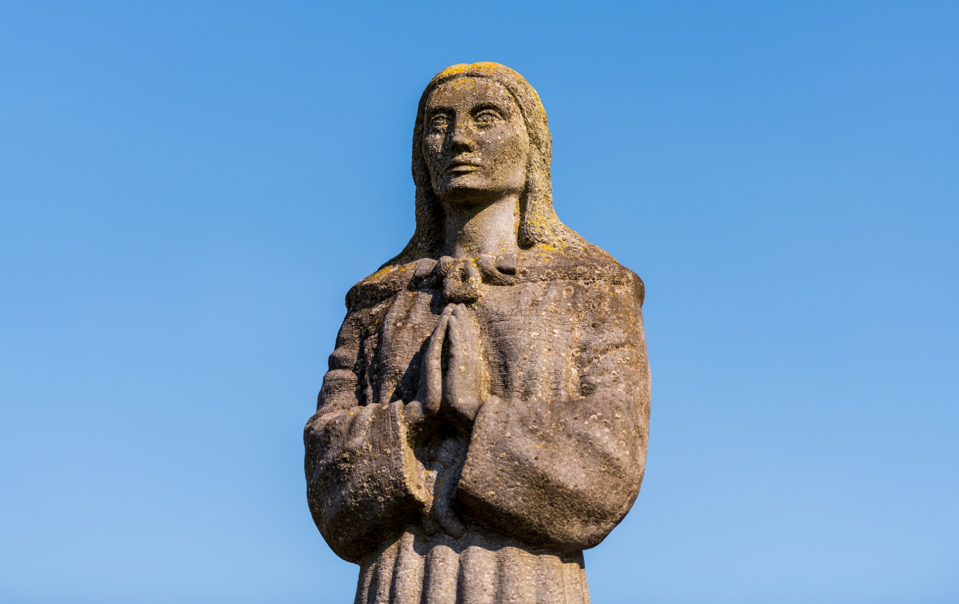 Close-up of St Brigid stone statue in Kildare town against a blue sky, with moss-covered detail and hands in prayer.