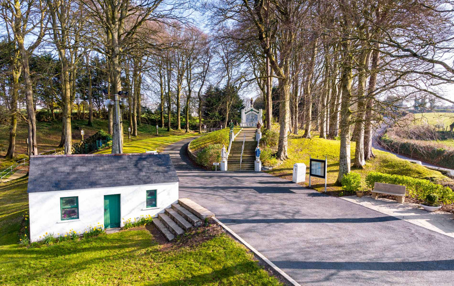 Pilgrimage site in Louth featuring a white cottage and tree-lined path leading to a religious shrine with statues of saints.
