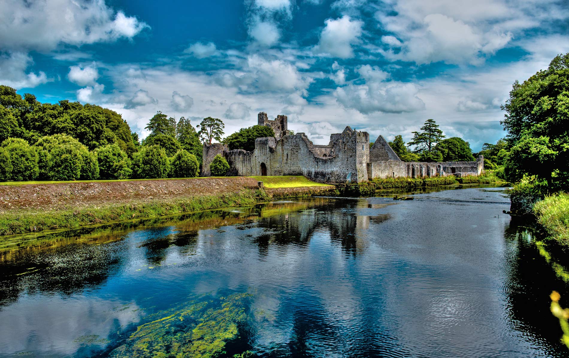 Castle ruins on the grounds of Adare Manor in County Limerick reflected in the river under a summer sky.