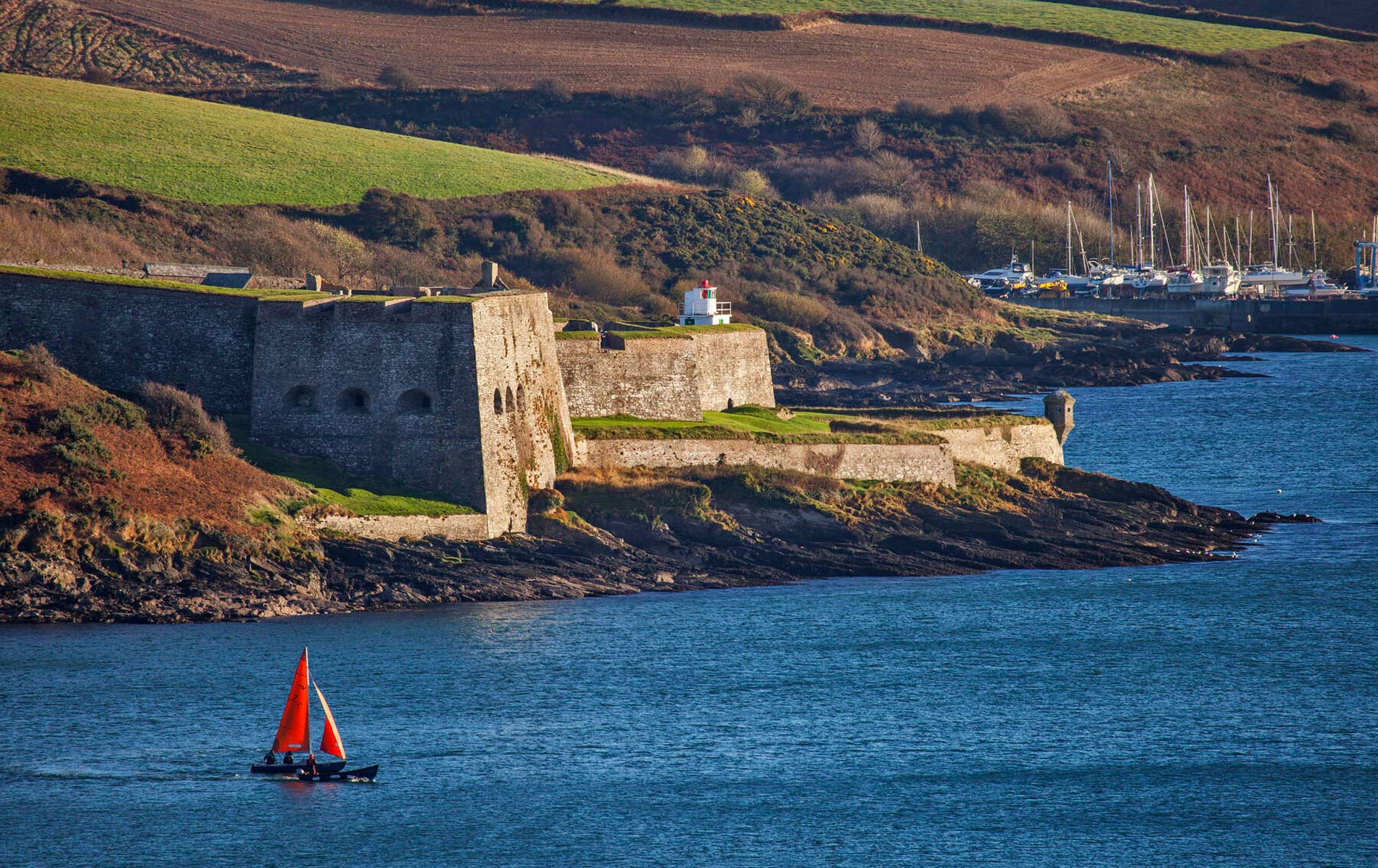 Coastal view of Charles Fort in Kinsale, County Cork, with a red-sailed boat in the harbour.