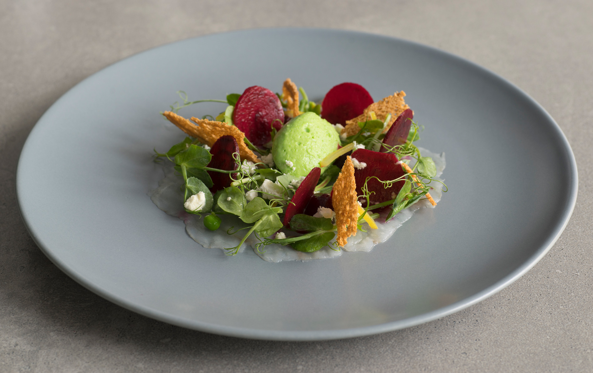Fine dining dish with fresh greens, beetroot, raspberries, and savoury crisps on a grey plate.