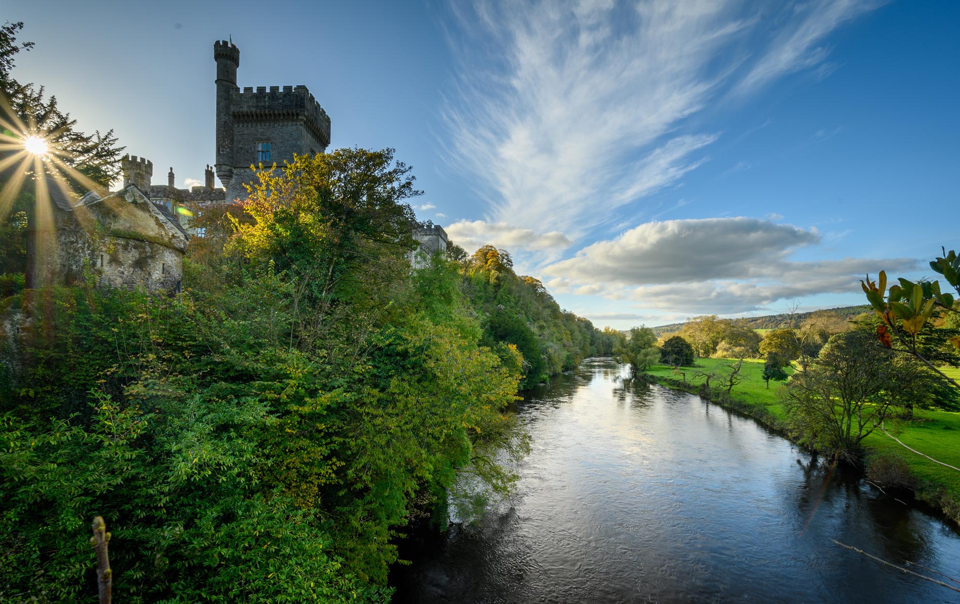 River flowing past Lismore Castle in County Waterford with sunlit stone tower and green meadows.
