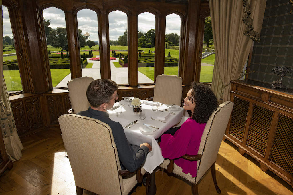 Pareja cenando en una sala con paneles de madera con vistas a jardines cuidados y jardines ornamentales en la finca Adare.