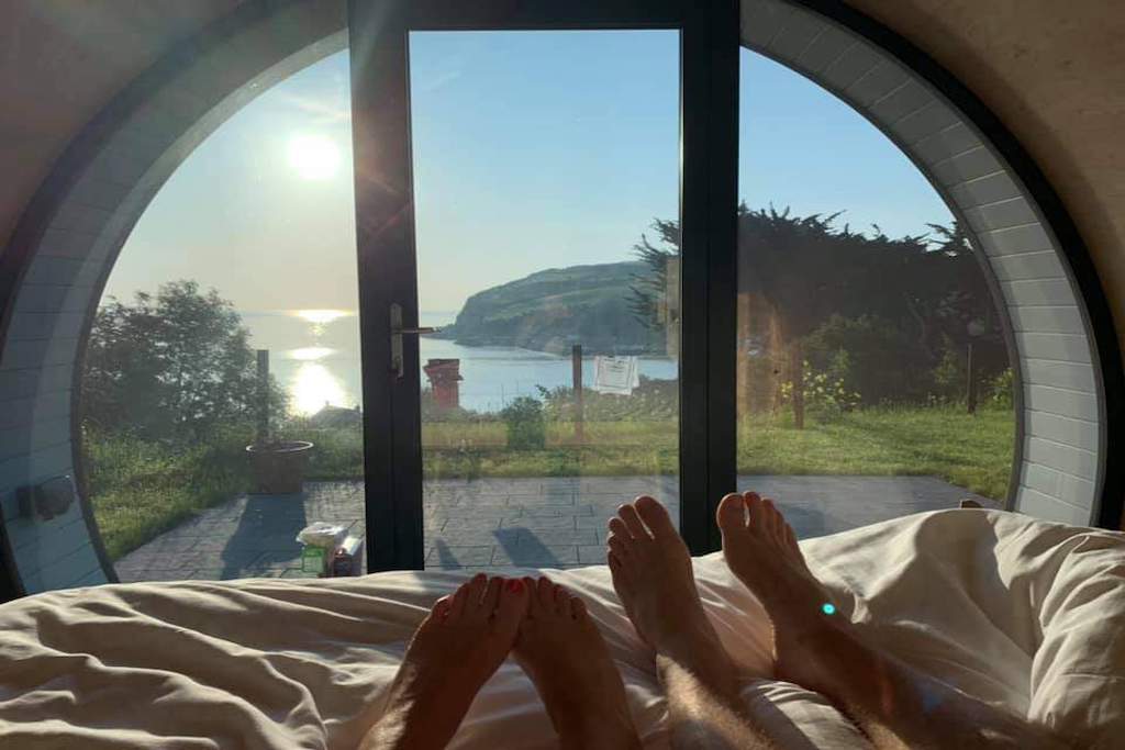 Dos personas relajándose en la cama dentro de una cápsula ecológica curvada, contemplando una vista costera iluminada por el sol en el Glenarm Castle, Antrim.