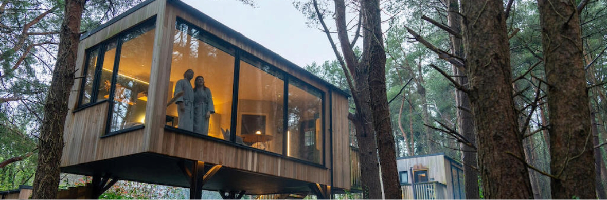 Cabane forestière de Burrenmore Nest avec de grandes fenêtres montrant un couple en peignoirs surplombant une forêt au crépuscule dans le comté de Londonderry.
