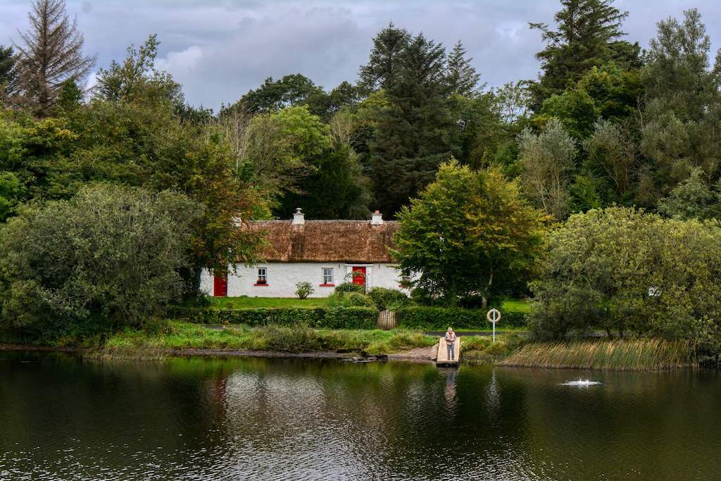 Thatched and whitewashed Keenaghan Cottage in County Fermanagh with a man fishing by the lake.