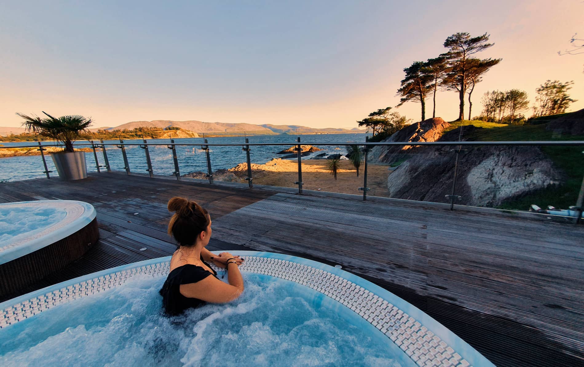 Woman sitting in a circular hot tub on a deck overlooking the sea and rocky coastline at Parknasilla Resort, Cork.