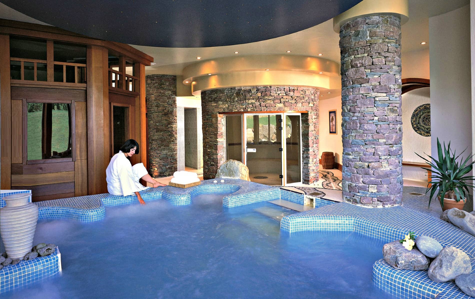 Spa guest in a robe dipping feet into an indoor thermal pool surrounded by curved stone pillars at Delphi Resort, Galway.