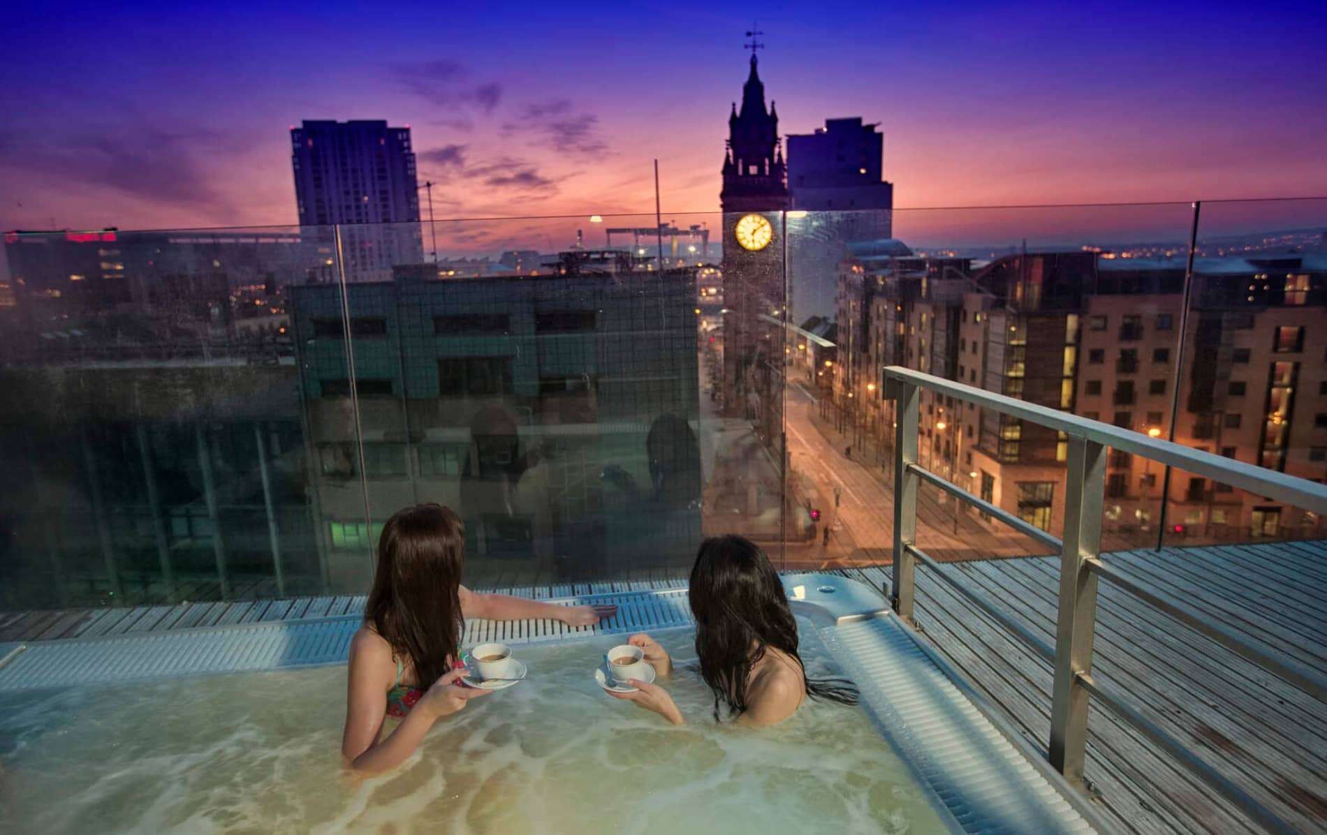 Two people in Merchant Hotel drinking coffee in a rooftop hot tub overlooking Belfast city skyline at twilight.