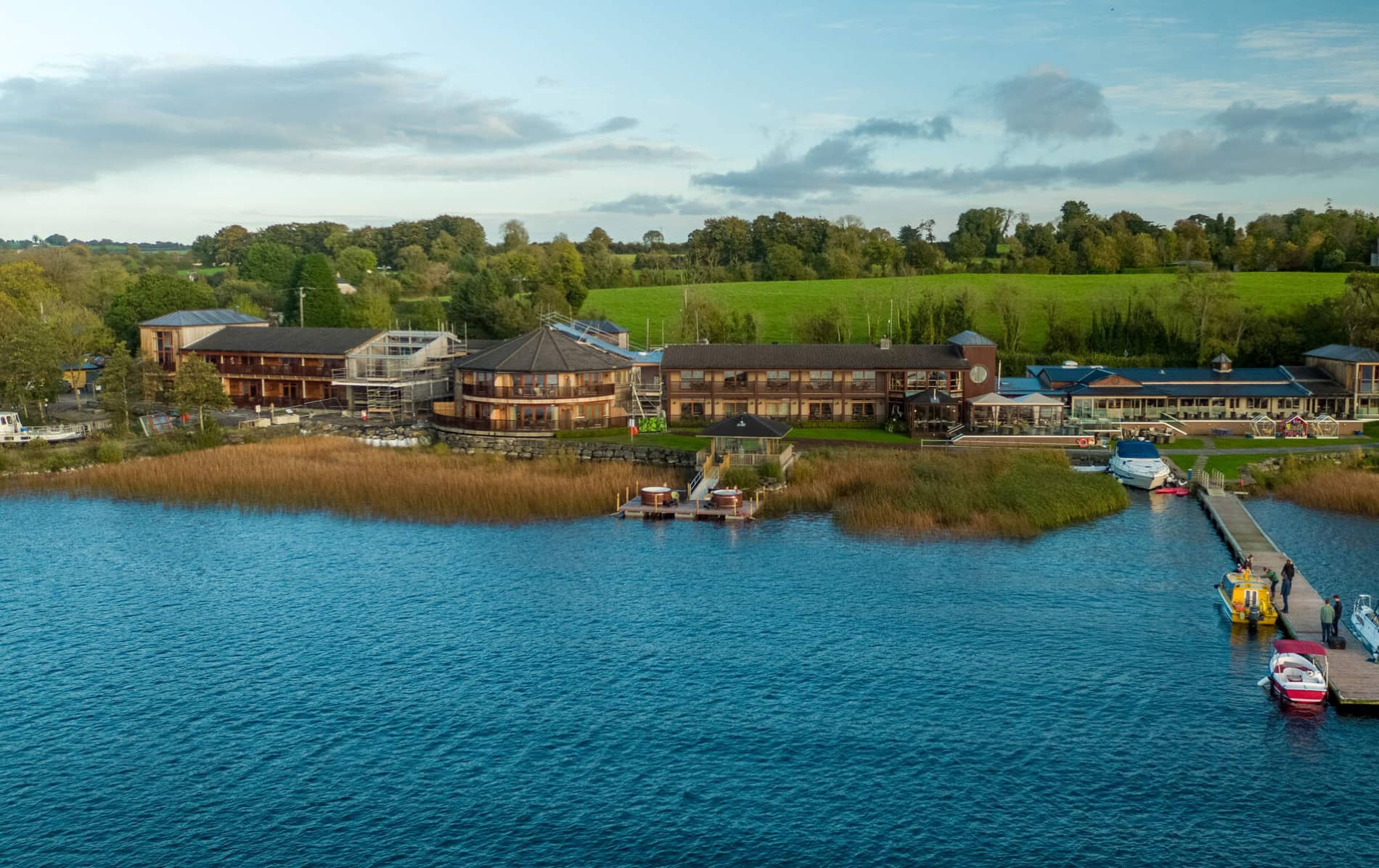 Aerial view of Wineport Lodge in Ireland with boats moored and outdoor hot tubs on wooden decking by the shore.