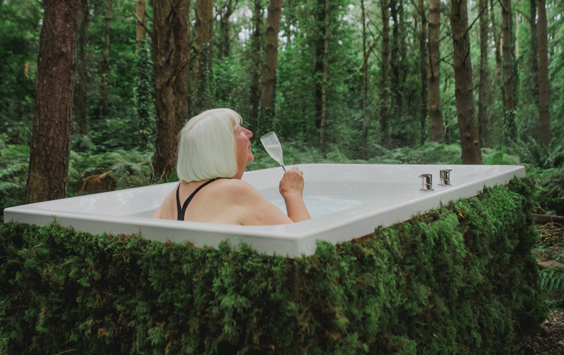 Older woman in a black swimsuit sipping sparkling wine in a moss-lined outdoor bath at Burrenmore Nest.
