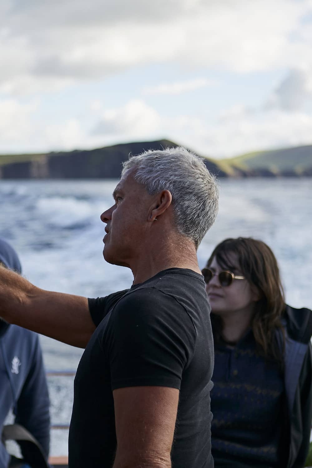 Un guía de vida marina masculina con cabello corto y gris señala hacia el horizonte mientras habla con los turistas en un barco frente a Dingle.