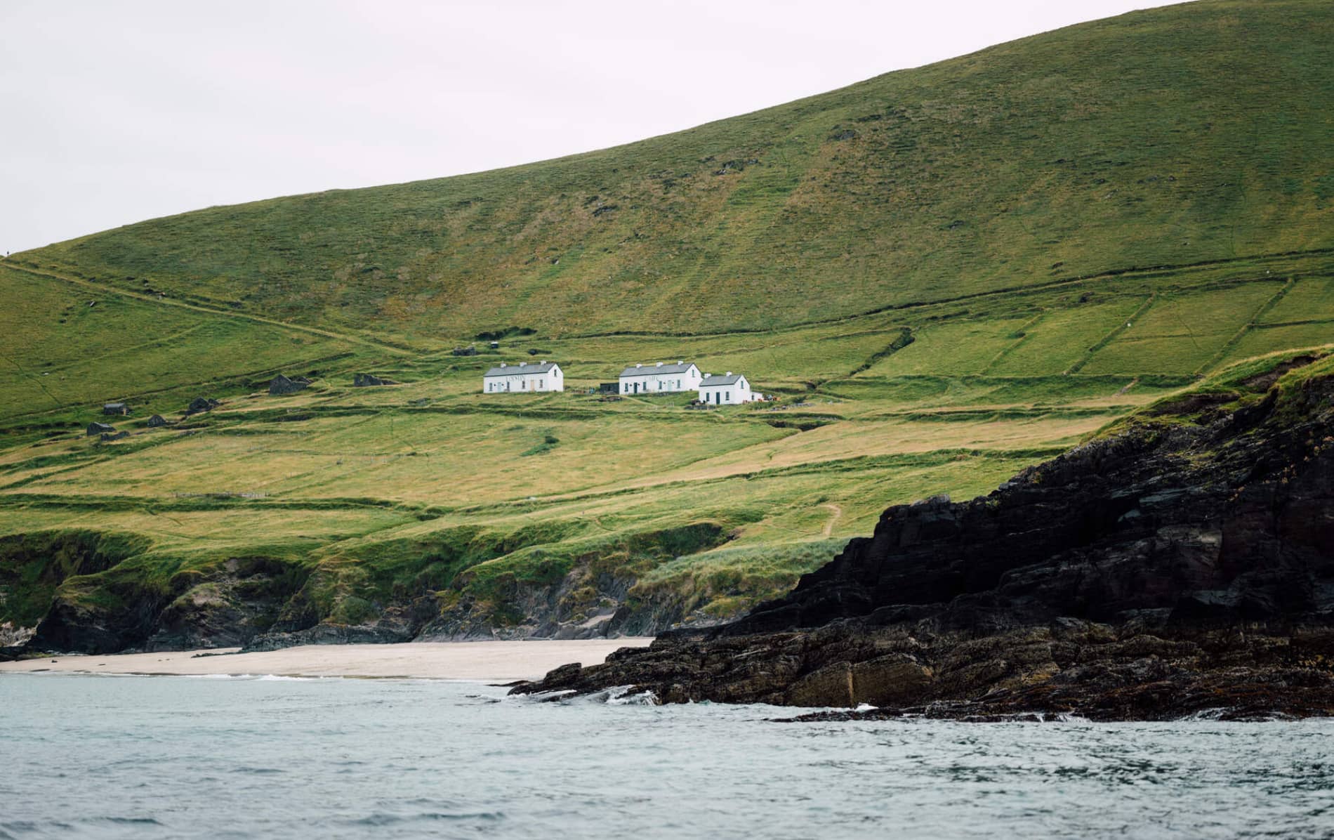 Cabañas encaladas esparcidas por lascolinas cubiertas de hierba de la islaGreat Blasket, con una playa de arena y Acantilados en la orilla.