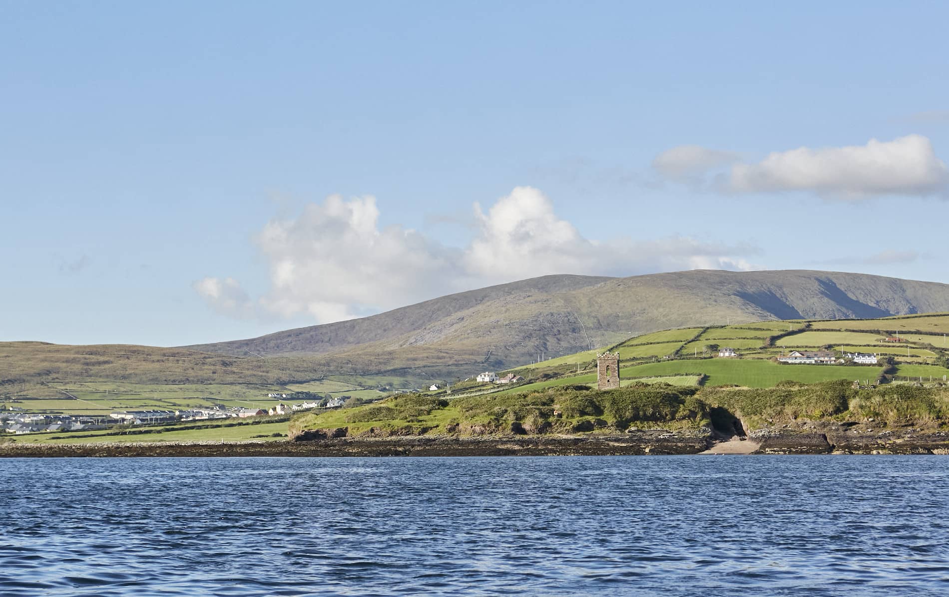 Vista del pueblo de Ballydavid con casas dispersas y una torre medieval contra las montañas de Slieve Mish bajo un cielo despejado.