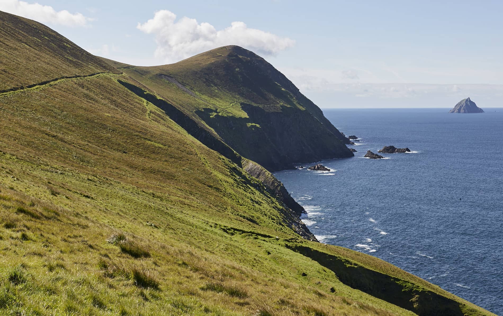 Acantilados verticales y laderas cubiertas de hierba de la Gran Isla Blasket que descienden hacia el mar, con una isla visible en el horizonte.