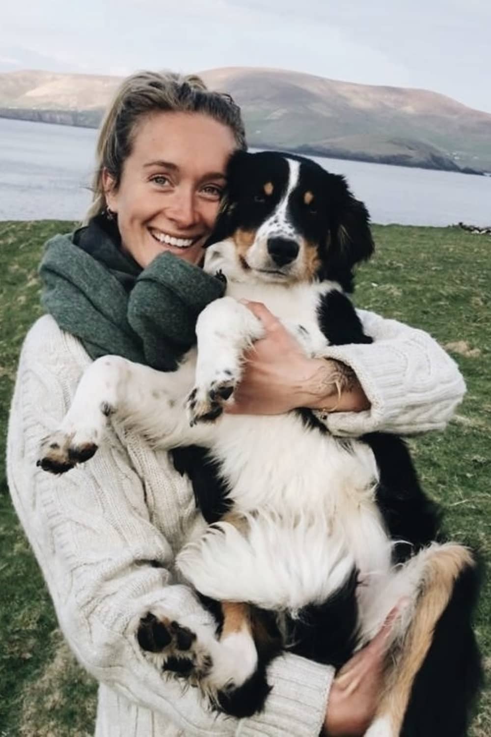 Mujer sonriente con un perro blanco y negro en la Gran Isla Blasket, azotada por el viento, con montañas y el océano al fondo.