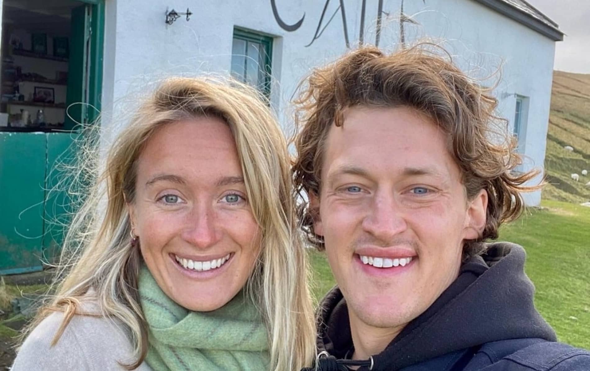 Pareja sonriente fuera de una casa blanca en la isla Great Blasket, tomando un selfie con el pelo azotado por el viento y colinas verdes detrás.