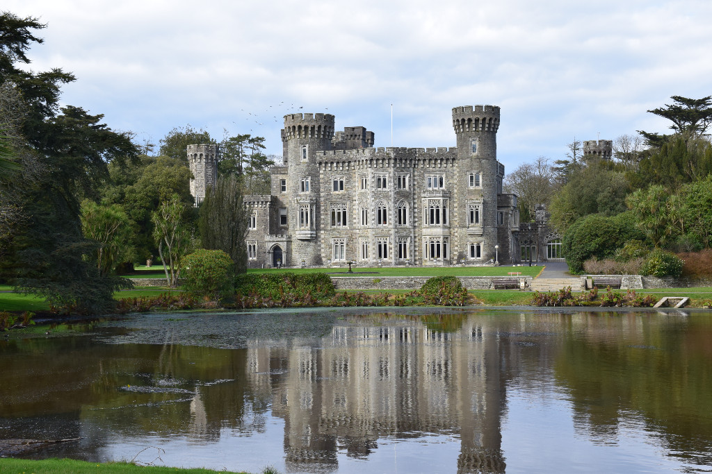 Johnstown Castle reflected in a pond, surrounded by formal gardens and woodland under a cloudy sky.
