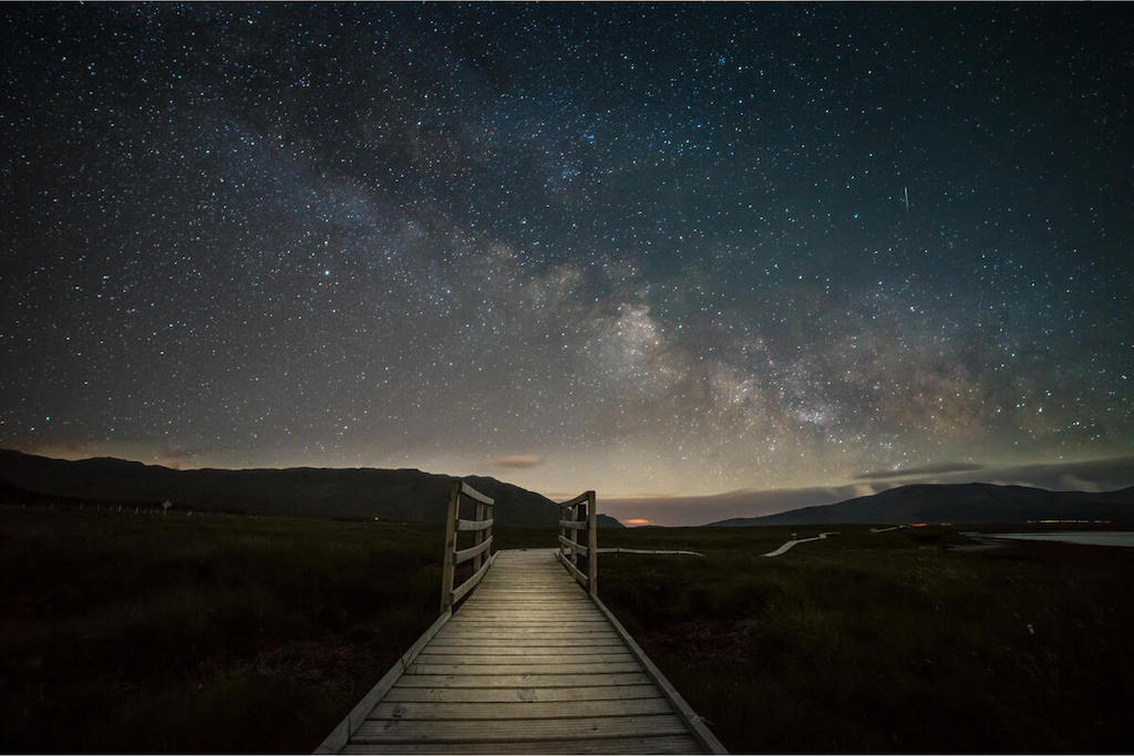 Houten promenade die naar een moeras in County Mayo leidt onder een heldere nachtelijke hemel vol sterren en de Melkweg.