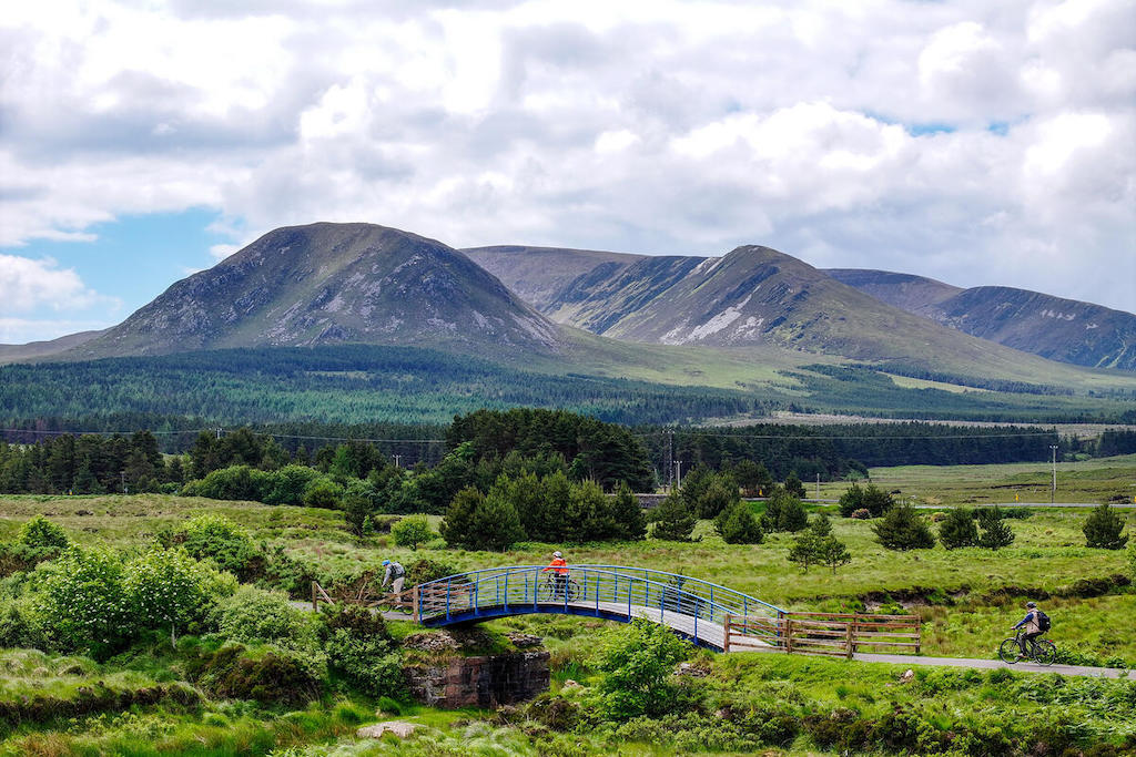 Fietsers die een brug oversteken op de Great Western Greenway in County Mayo, met groene velden en bergen op de achtergrond.