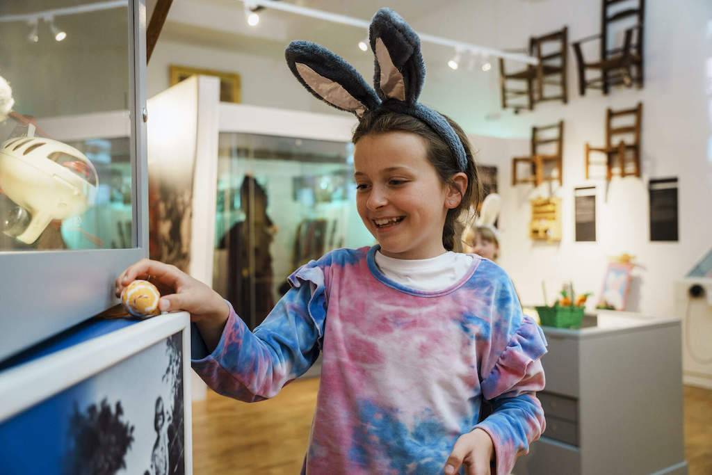 Smiling child wearing bunny ears headband during an Easter egg hunt in a brightly lit Tipperary Museum of Hidden History.
