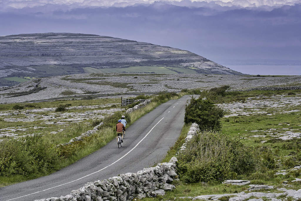 Two cyclists riding along a quiet road through the rocky limestone landscape of the Burren, County Clare.