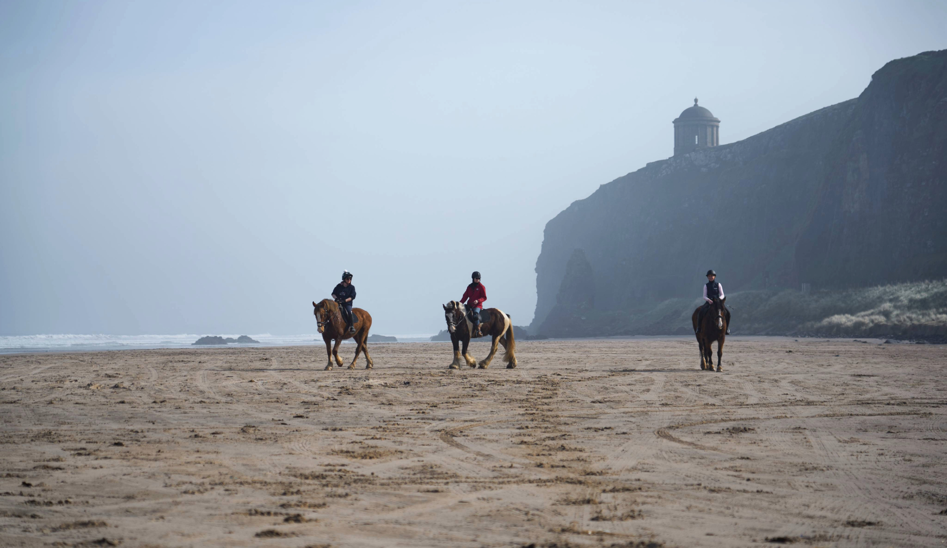 Three horse riders cross Downhill Beach, County Londonderry, with Mussenden Temple perched on the cliffs above in the mist.