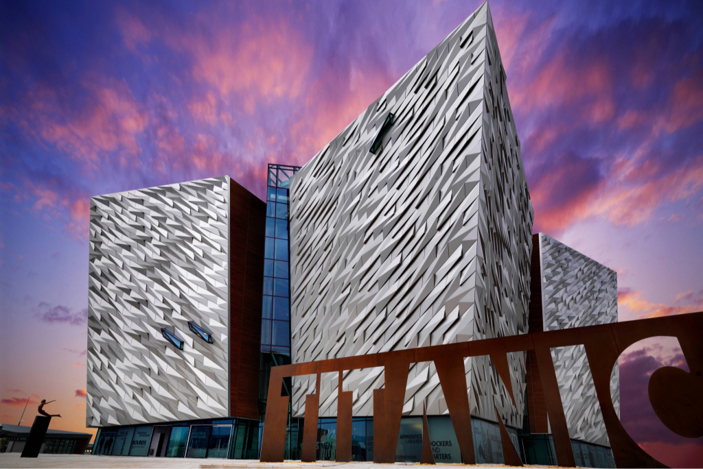 Titanic Belfast's angular, silver-clad exterior beneath a pink and purple sunset sky, with large TITANIC lettering in front.