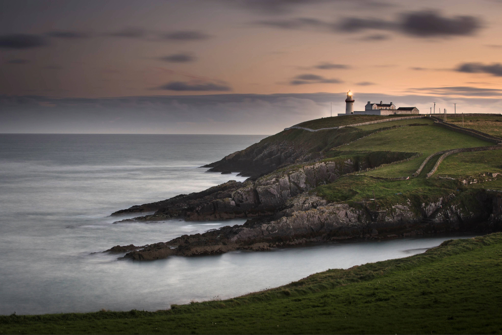 Galley Head Lighthouse on a grassy headland in County Cork, overlooking the Atlantic Ocean and rocky coastline at dusk.