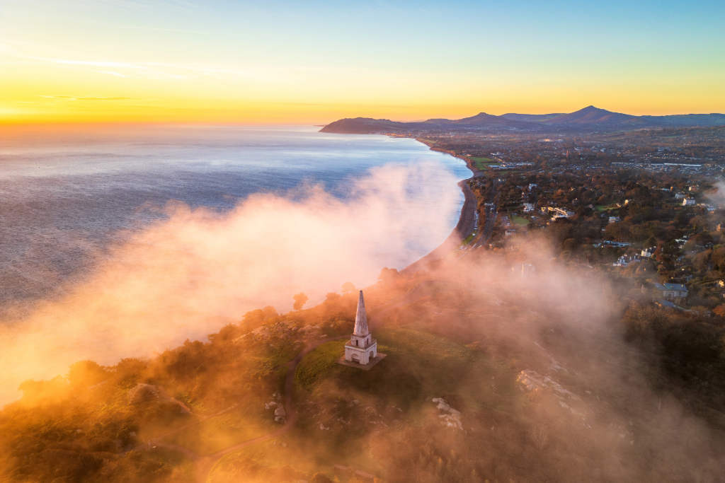 The obelisk on Killiney Hill in County Dublin overlooks the coastline and Irish Sea at sunrise, with mist drifting over the trees.
