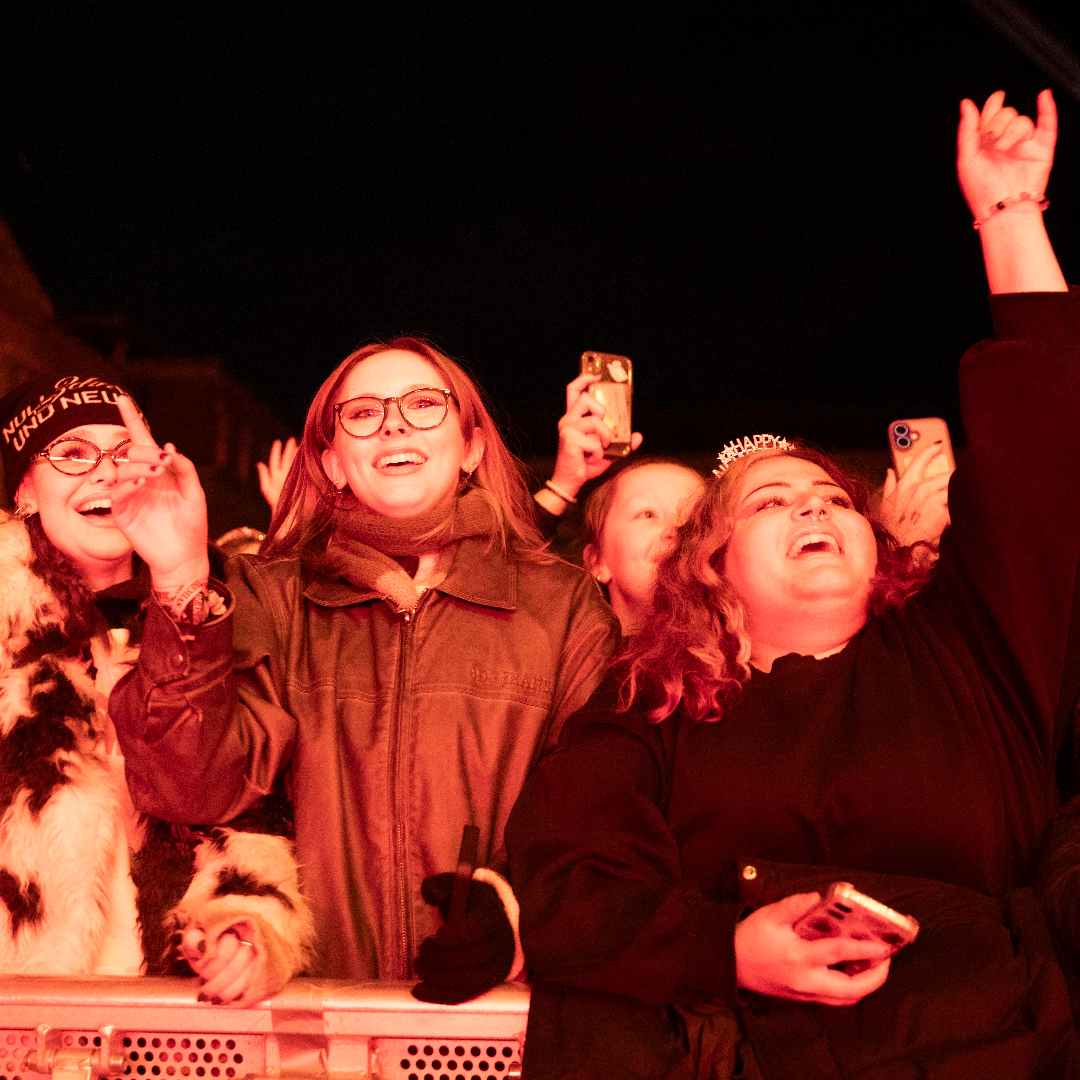 Festivalgoers cheering, smiling and raising their hands behind a crowd barrier at New Year's Festival Dublin, illuminated by bright red stage lighting.