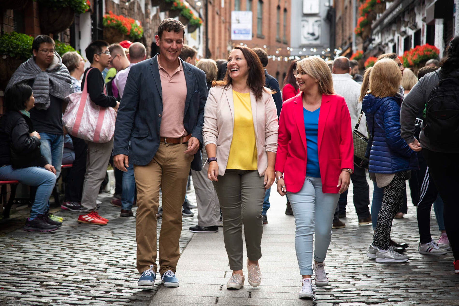 Friends walking along a busy cobbled street in Belfast's Cathedral Quarter filled with people.