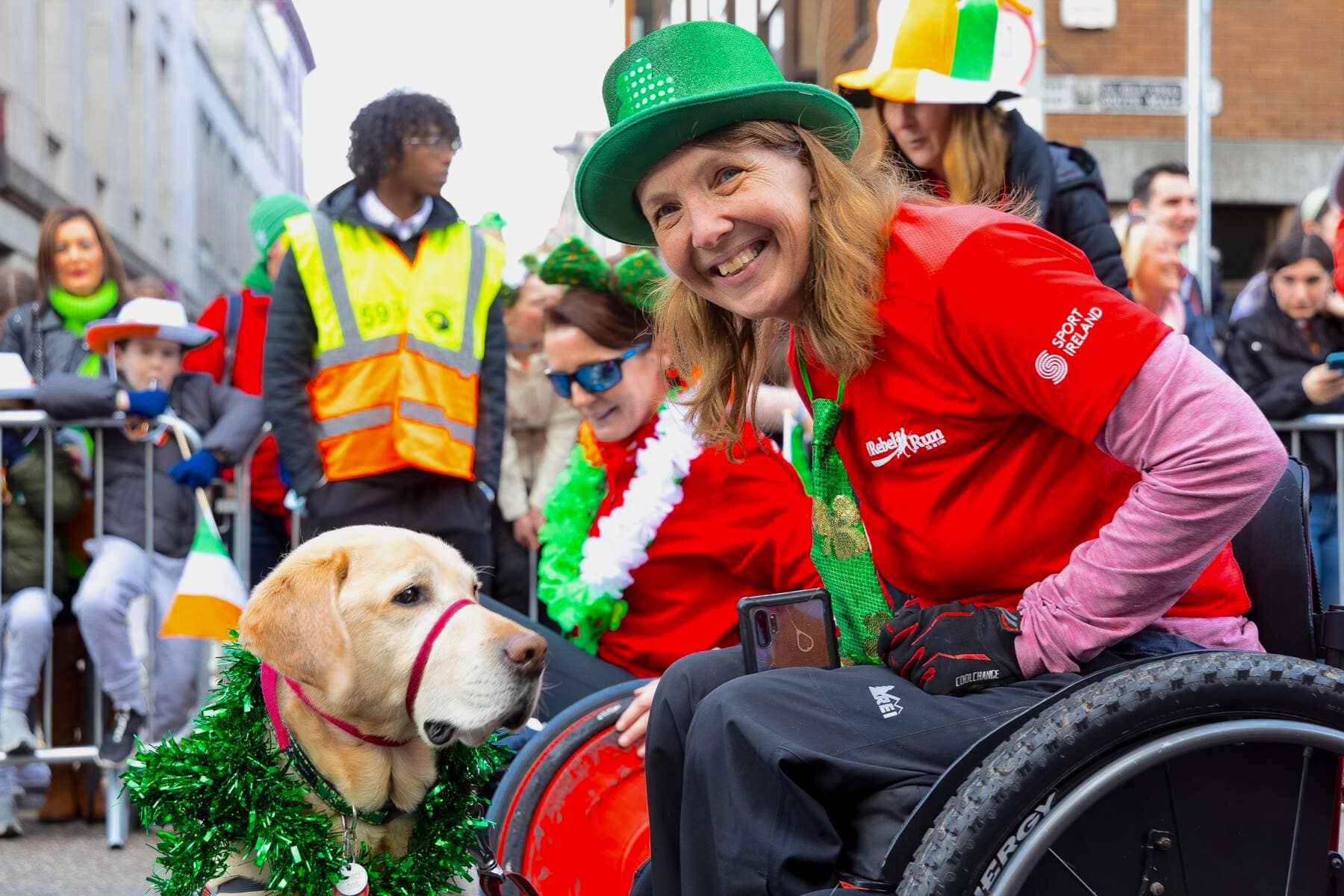 Une femme souriante en fauteuil roulant portant un chapeau vert pose avec son chien-guide lors d'un défilé de la Saint Patrick à Cork.