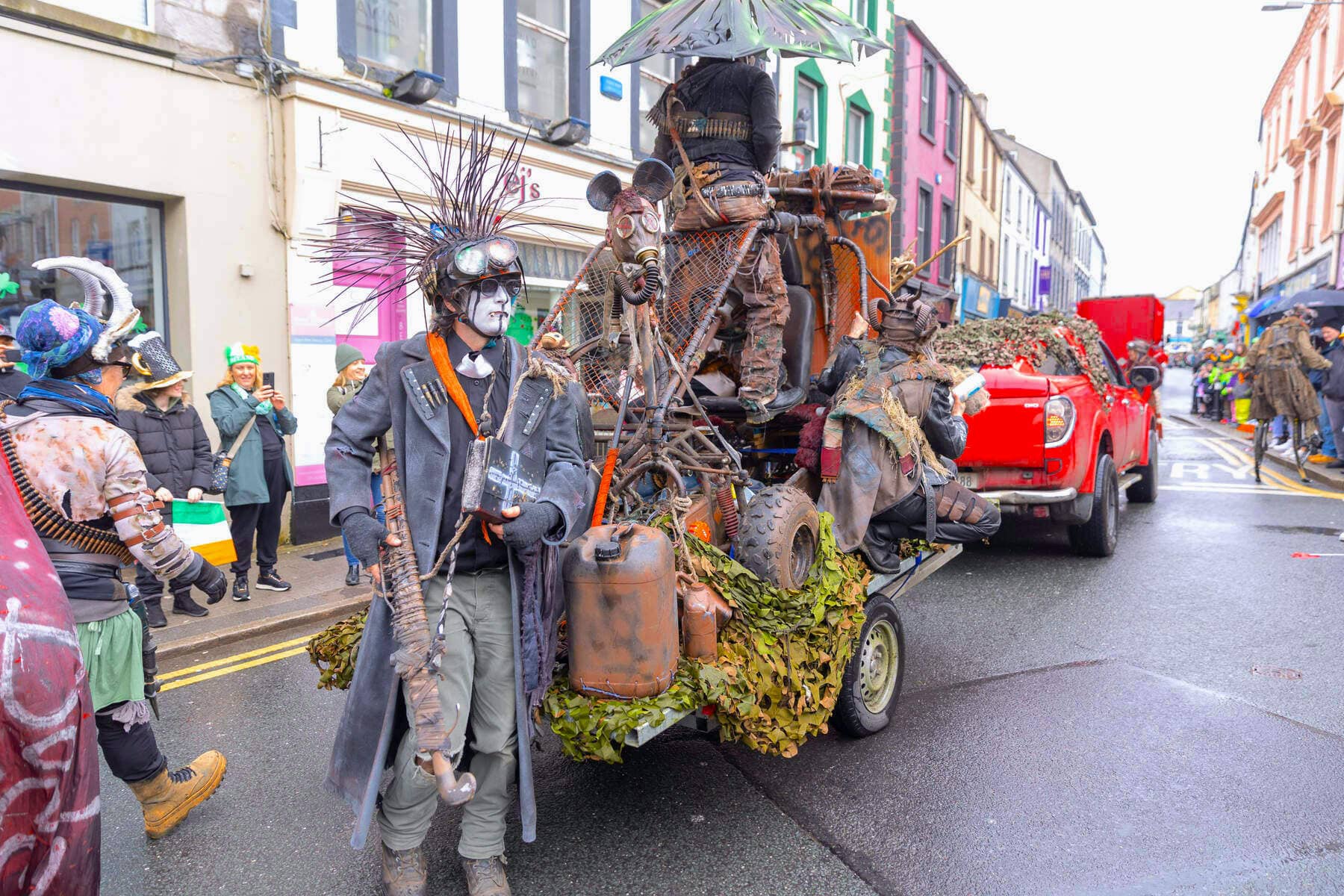 Des artistes de parade en costumes spectaculaires avec des accessoires défilent à côté d'une charrette décorée dans une rue de Sligo.