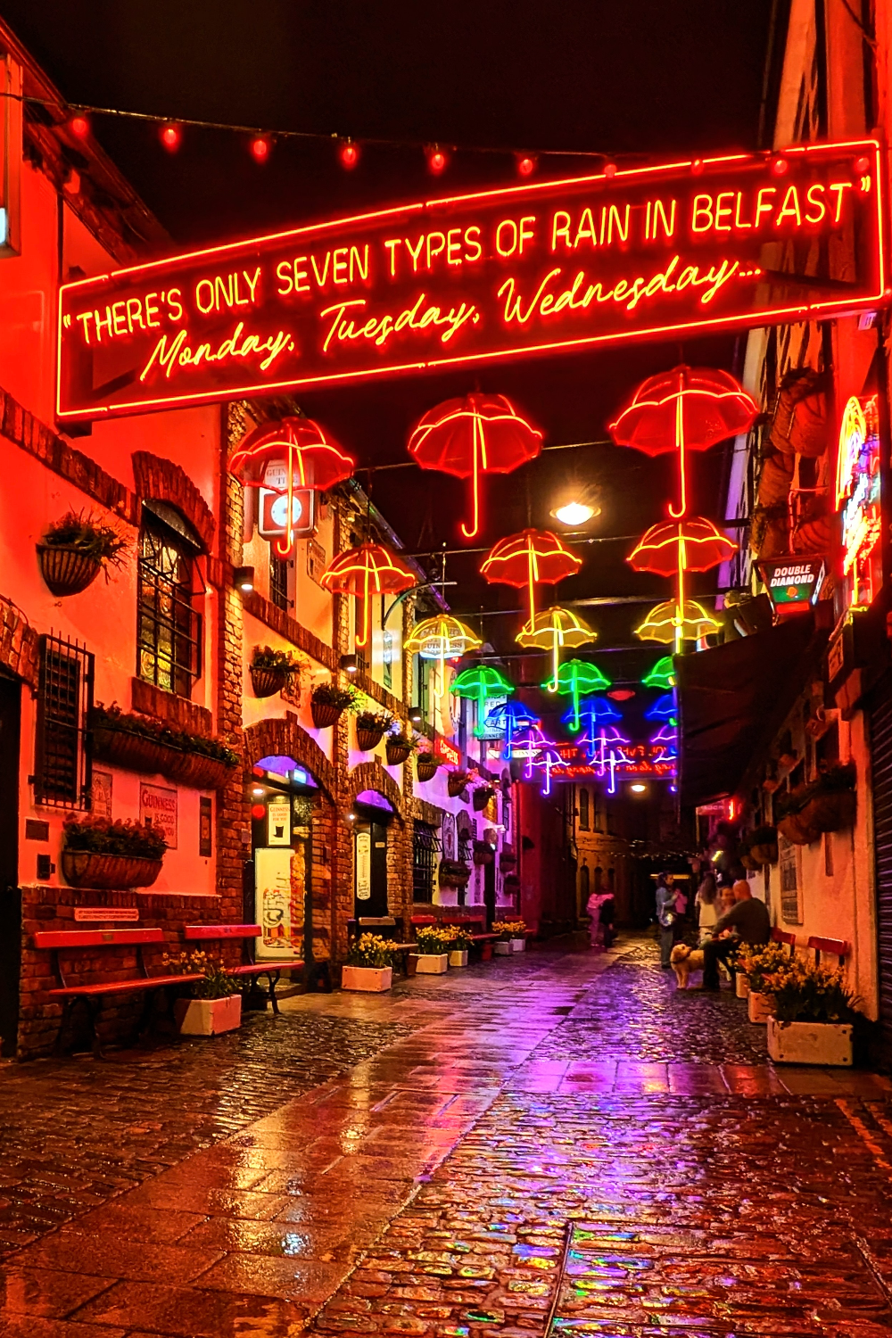 The Duke of York in Belfast at night, with colourful neon umbrellas reflected on the wet cobbled street.