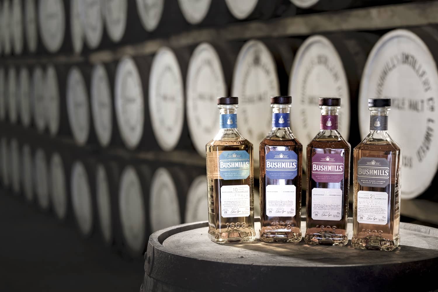 Bottles of Bushmills Irish whiskey displayed on a barrel in front of rows of ageing whiskey casks.