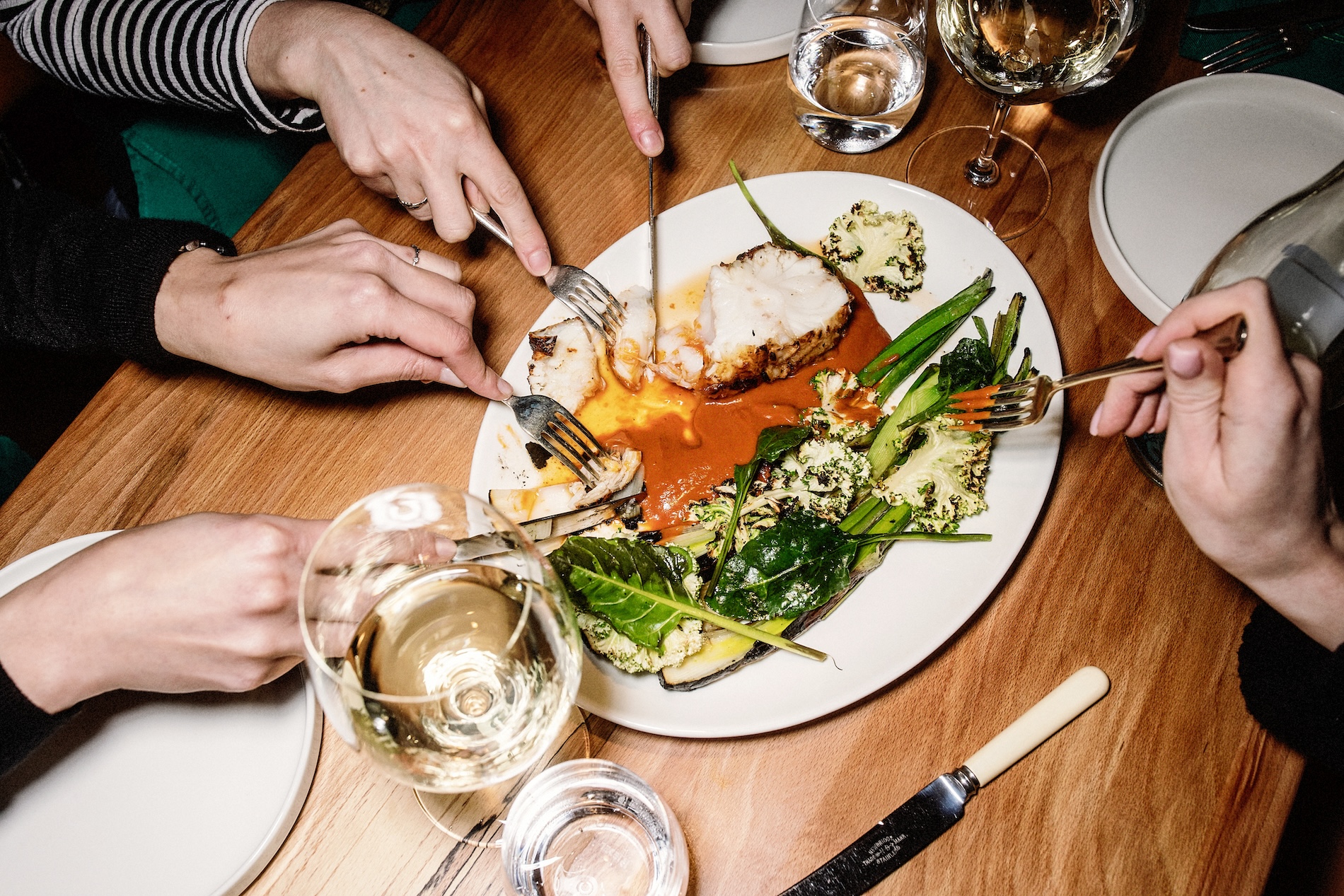 Diners share a seafood dish with grilled vegetables and wine at a wooden table.